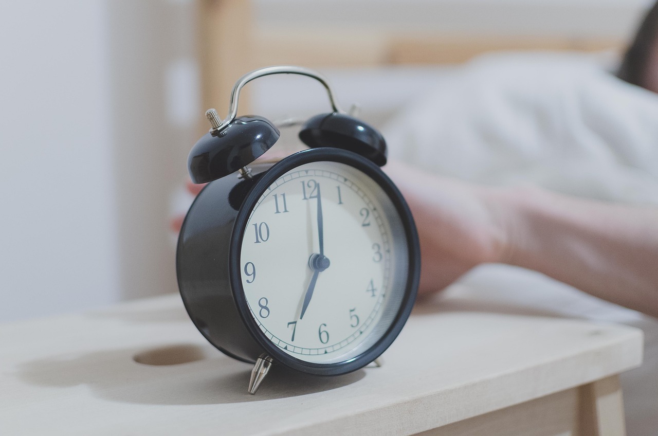 A classic black twin-bell alarm clock on a wooden nightstand set for seven, with a sleepy arm reaching toward it from the bed