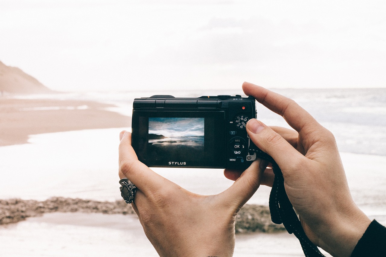 Two hands holding a compact point-and-shoot camera at the edge of a beach, framing the shoreline on its rear screen