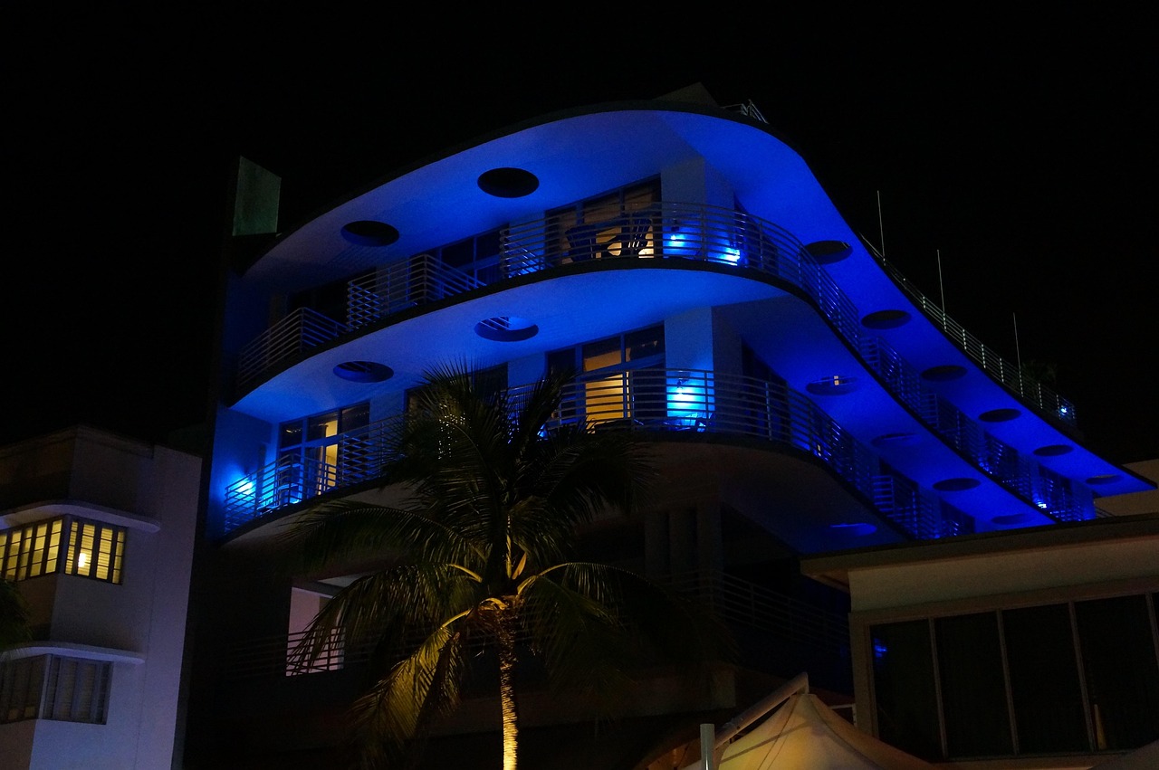 An Art Deco hotel in Miami Beach lit up in electric blue at night with a palm tree in front, the real-world mood behind Vice City