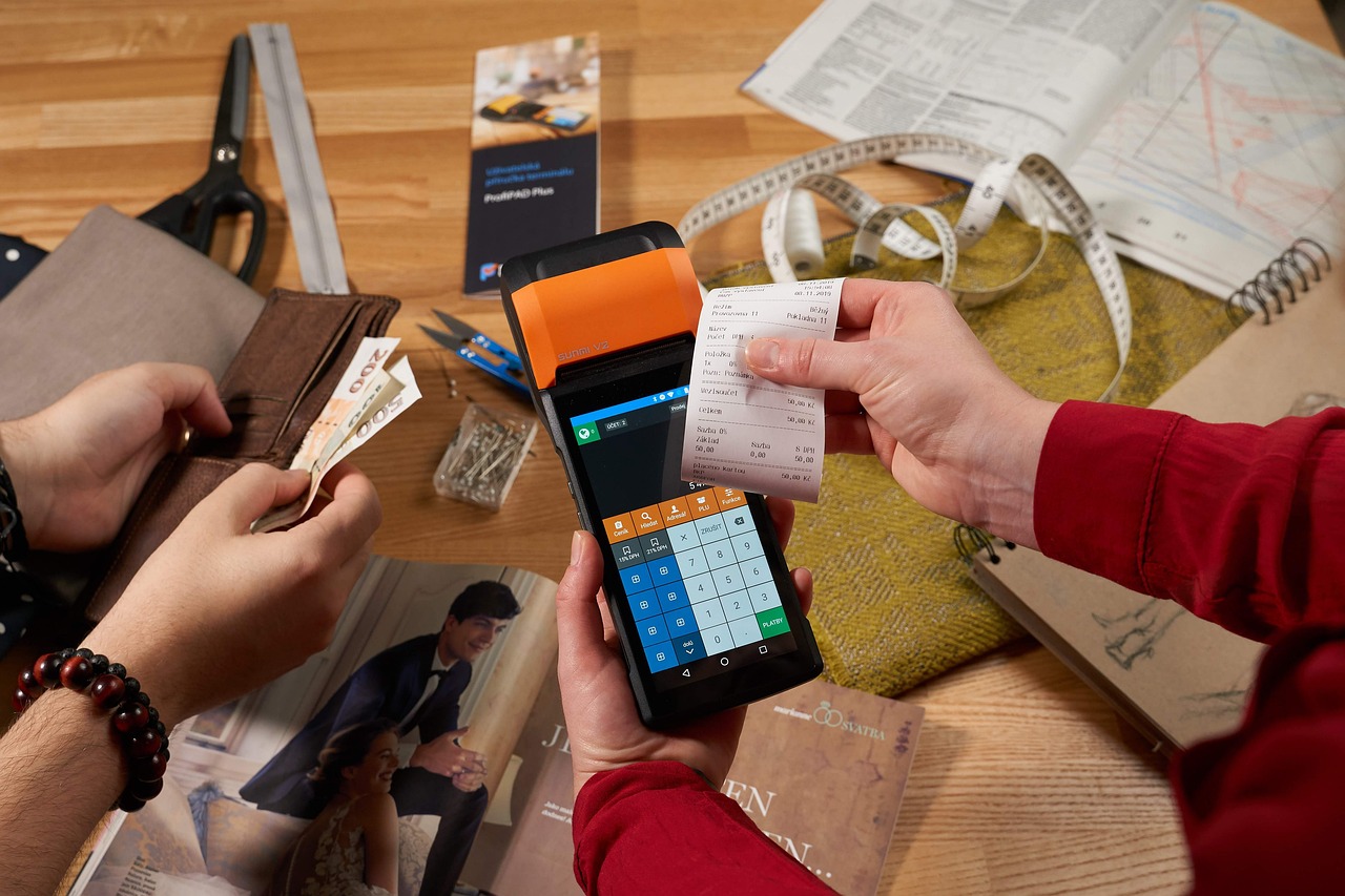 A handheld card terminal printing a receipt while someone pulls cash from a wallet, the moment a laptop price actually lands