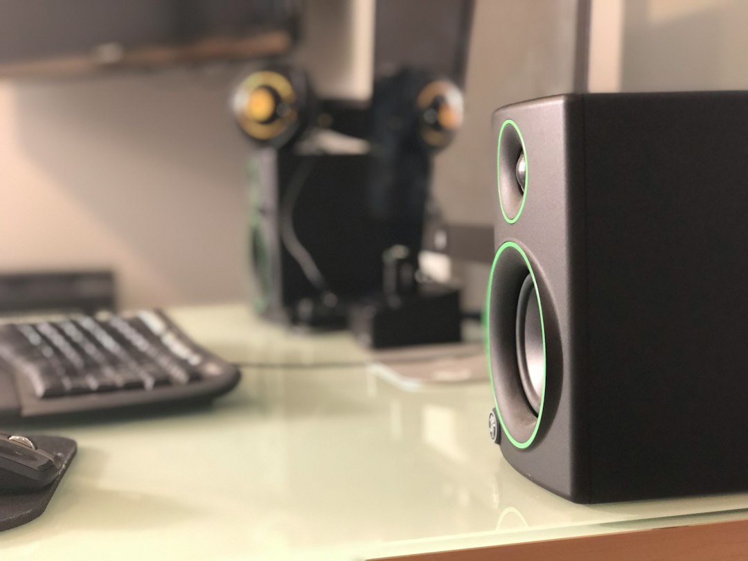 Black nearfield monitors with green rings on a glass desk, headphones blurred behind them