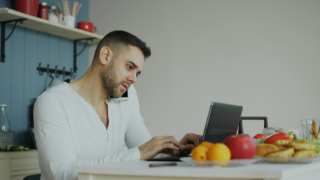 A man on a phone call while typing on a laptop at a kitchen table with fruit and pastries nearby