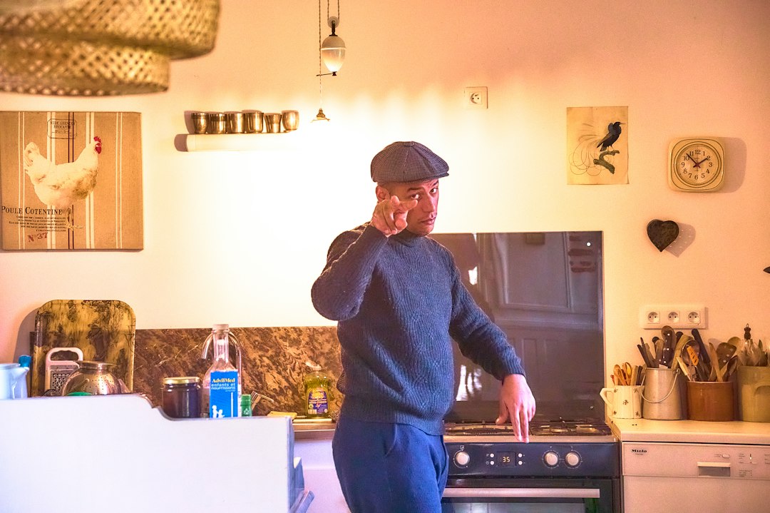 Older man in a flat cap pointing sternly across his kitchen