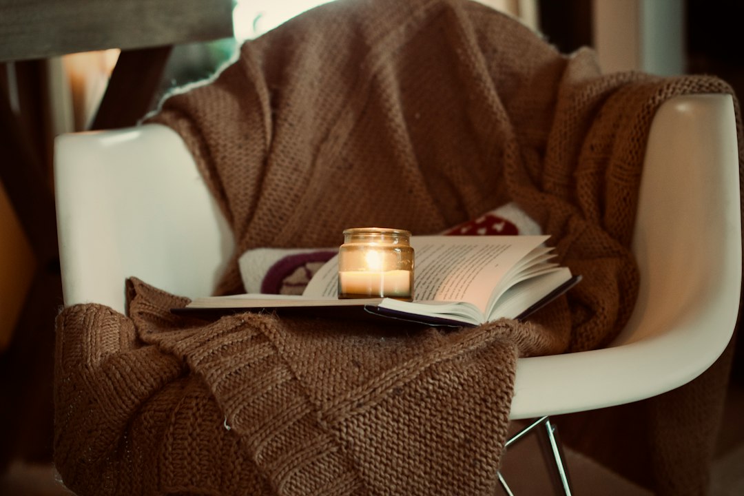 An open book and a lit candle in a jar resting on a chunky knit blanket draped over a chair, a cozy evening without screens