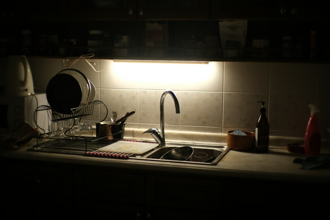 A dim kitchen counter and sink lit by a single under-cabinet light at night