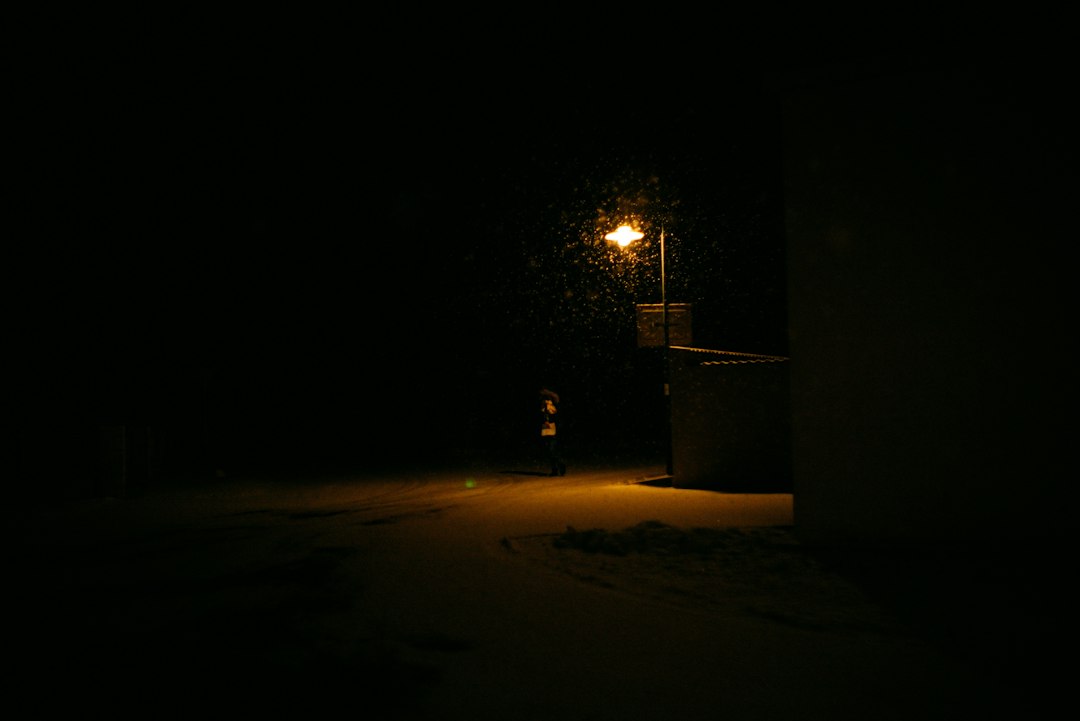 A lone figure walking a snowy street under a glowing street lamp at night