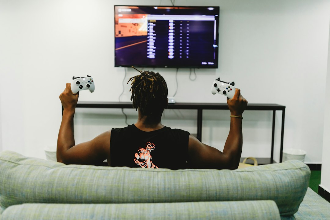 A person on a couch facing a wall-mounted TV raises two white game controllers in celebration