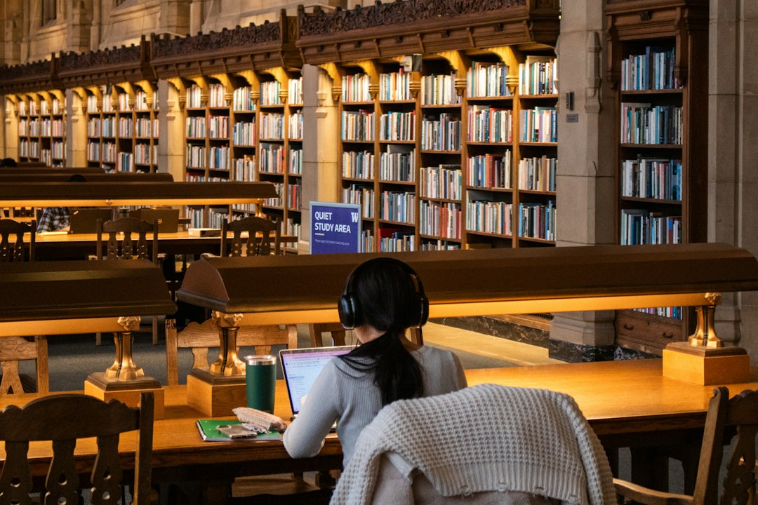 A student wearing headphones works on a laptop at a long table in a grand library